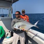 Captain John smiling on a boat holds a large, shimmering Pompano fish with the clear blue sea behind him, captured on one of the best fishing charters in Key West, with another person in the background admiring the catch.