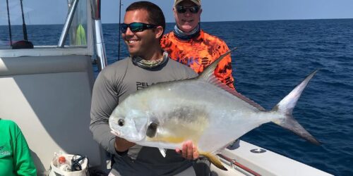 Captain John smiling on a boat holds a large, shimmering Pompano fish with the clear blue sea behind him, captured on one of the best fishing charters in Key West, with another person in the background admiring the catch.