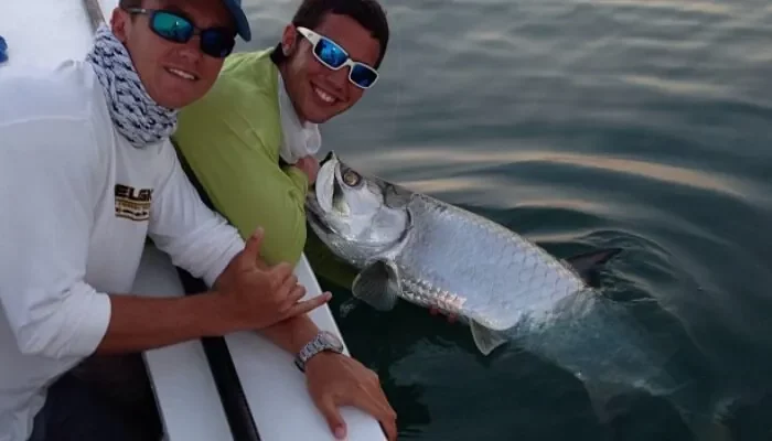 Two fishermen in sunglasses and caps are seen smiling beside a large tarpon at sunset, illustrating the rewarding experience of tarpon fishing in Key West.